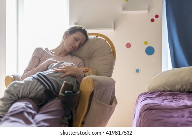 Tired Young Mother Resting Or Napping On A Rocking Chair With Baby Sleeping In Her Lap Inside A Nursery Room At Day.