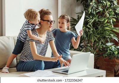 Tired Young Mother Sitting On Sofa And Working With Laptop And Documents While Little Kids Having Fun And Making Noise
