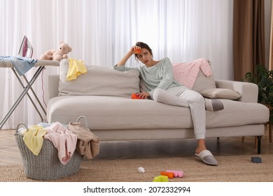 Tired Young Mother Sitting On Sofa In Messy Living Room