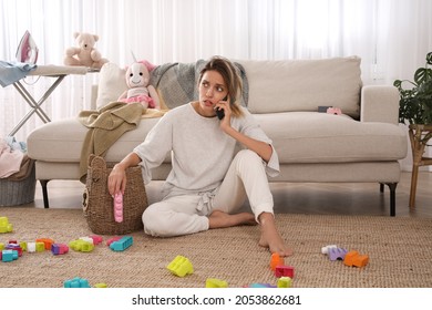 Tired young mother talking on phone in messy living room
