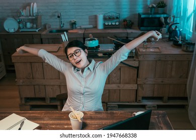 Tired Young Office Lady Employee Sitting At Dark Home Kitchen In Night And Raising Arms Stretching Body After Long Time Working. Female Enjoy Eating Unhealthy Bedtime Snack Instant Noodles And Relax