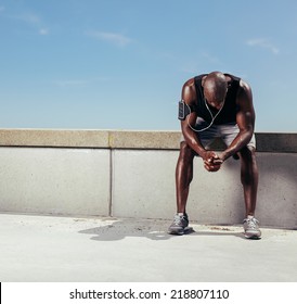Tired young runner leaning over to catch his breath. African man sitting on a wall relaxing after fitness workout outdoors.
