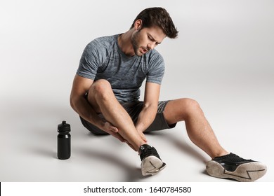 Tired young sportsman resting on a floor isolated over gray background, having ankle pain