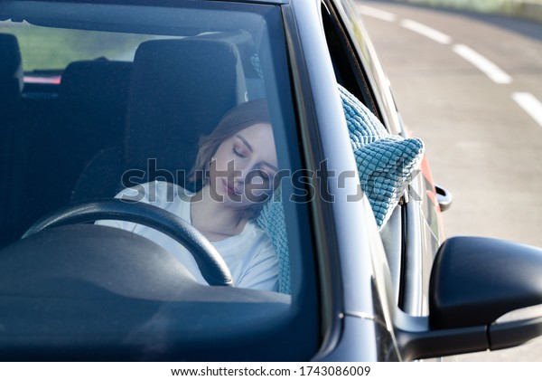 Tired young woman driver asleep on pillow on steering\
wheel, resting after long hours driving a car. Fatigue. Sleep\
deprivation. 