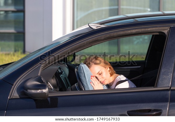 Tired young woman driver asleep on pillow on steering\
wheel, resting after long hours driving a car. Fatigue. Sleep\
deprivation. 