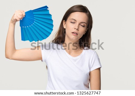 Tired young woman feel overheated suffering from heat stroke high temperature sweating problem, sweaty girl holding waving fan cooling in hot summer weather isolated on white grey studio background