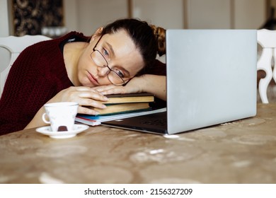Tired young woman in glasses lying down on books and looking away near laptop at desk in home room. Tiredness from monotonous study and computer overwork. Lack of energy motivation concept