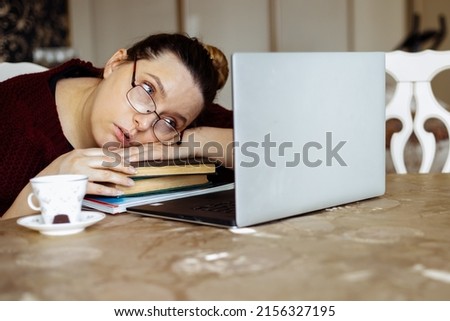 Tired young woman in glasses lying down on books and looking at laptop screen at desk in home room. Tiredness from monotonous study and computer overwork. Lack of energy motivation concept