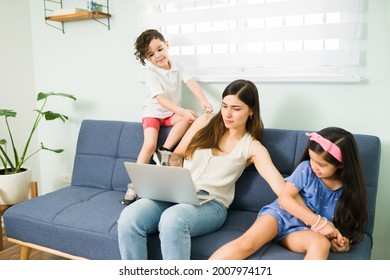 Tired young woman trying to work with a laptop at home. Playful children bothering their working mom and trying to play with her 