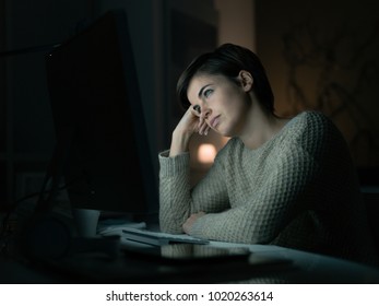 Tired young woman working with her computer late at night, she is leaning on her hand and staring at the computer screen