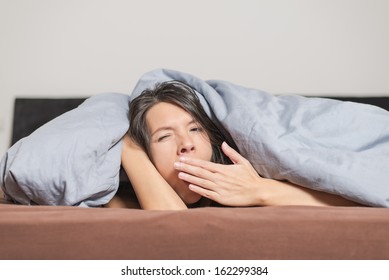 Tired Young Woman Yawning Under A Duvet Holding Her Hand In Front Of Her Mouth As She Spends A Lazy Day Relaxing At Home