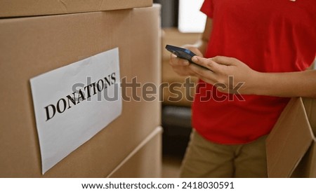 Tireless female volunteer's hands, a resilient hispanic woman, dutifully using her smartphone at heartwarming charity center amid boxes ready for donation packaging.