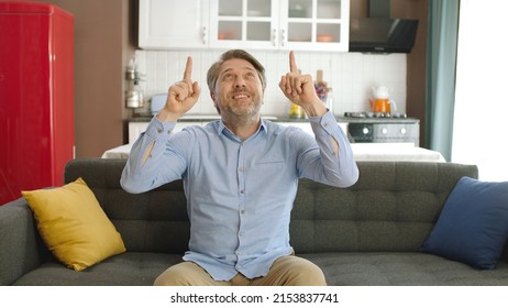 Title: Young man pointing fingers top of screen at camera while sitting on comfortable sofa in his apartment. Creative people can put whatever they want where the man points.