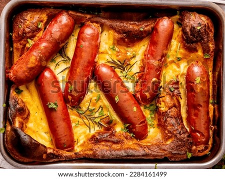 Toad in the hole, close-up top view, horizontal image with fresh parsley and rosemary sprigs. Baked sausages in Yorkshire pudding