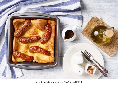 Toad In The Hole Of Yorkshire Pudding With Roasted Sausages Served On A Square Cake Pan With Onion Gravy, Plate And Cutlery On A White Wooden Background, Horizontal View