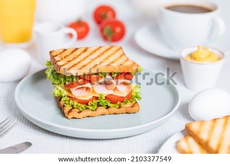 Toast sandwich with gouda cheese and turkey ham filled with tomato and lettuce on a bright background with ingredients in blurry background. Served with coffee and orange juice and eggs