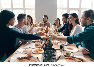 Toast to us! Group of young people in casual wear toasting each other and smiling while having a dinner party indoors