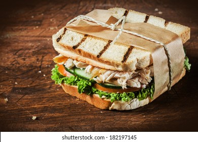 Toasted Shredded Chicken Breast And Salad Sandwich Served On A Rustic Wooden Table Wrapped In A Brown Paper Wrapper Tied With String With Vignetting And Copyspace