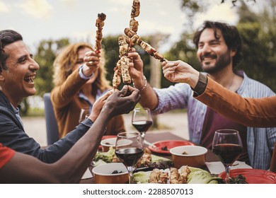 Toasting with Skewers - Group of people in their 30s and 40s at a garden table, holding meat skewers and toasting. Mixed ethnicity. Blurred trees and sunset in the background. - Shutterstock ID 2288507013