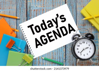 Today's Agenda text on paper from a notepad on a blue wooden background near colored notepads and an alarm clock - Shutterstock ID 2141373979