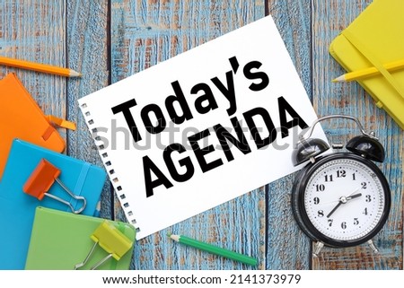 Today's Agenda text on paper from a notepad on a blue wooden background near colored notepads and an alarm clock