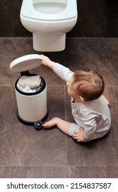 Toddler Baby Boy Is Playing In The Toilet Room With A Trash Can. Child Plays On A Brown Floor In A Beige Bathroom With A Trash Can Lid