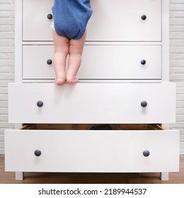 Toddler baby climbed up on the open chest of drawers. Child boy stood on a tall drawer of a white cabinet. Kid age one year