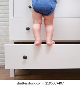 Toddler Baby Climbed Up On The Open Chest Of Drawers. Child Boy Stood On A Tall Drawer Of A White Cabinet. Kid Age One Year