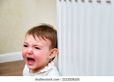 Toddler Baby Crying At The Radiator, Tearful Child Face Close-up. White Radiator For Heating The Home Room And Baby