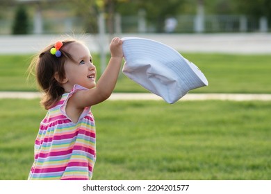 Toddler Baby Girl Wears Bright Striped Summer Bodysuit, Take Off And Holds Out Her White Panama Bucket Hat. Little Kid Child Walks In Summer Public Park