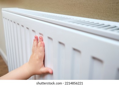 Toddler Baby Holding On To The Radiator, Child Hand On The Heating System Close-up. White Radiator For Heating The Home Room And Children's Hand