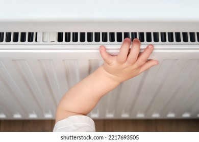 Toddler Baby Holding On To The Radiator, Child Hand On The Heating System Close-up. White Radiator For Heating The Home Room And Children's Hand