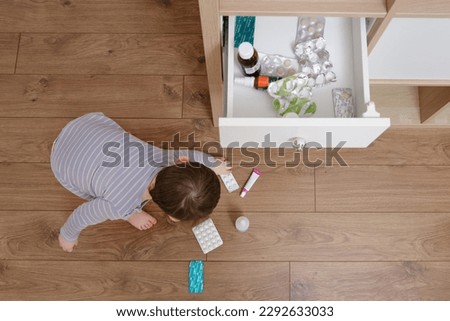 Toddler baby opened the cabinet drawer with pills and medicine. Child boy holding a pack of pills in the home living room. Kid age one year nine months