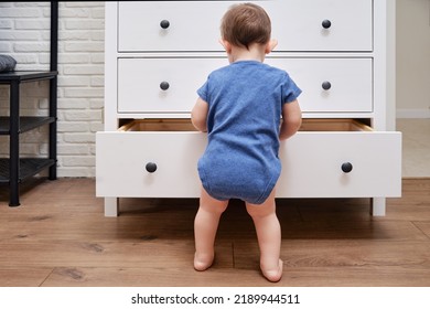 Toddler Baby Opens A Chest Of Drawers. Child Boy Reaches Into An Open Drawer Of A White Cabinet. Kid Age One Year