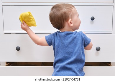 Toddler Baby Opens A Chest Of Drawers. Child Boy Reaches Into An Open Drawer Of A White Cabinet. Kid Age One Year