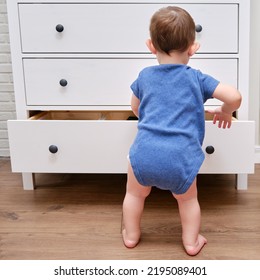 Toddler Baby Opens A Chest Of Drawers. Child Boy Reaches Into An Open Drawer Of A White Cabinet. Kid Age One Year