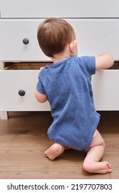 Toddler Baby Opens A Chest Of Drawers. Child Boy Reaches Into An Open Drawer Of A White Cabinet. Kid Age One Year