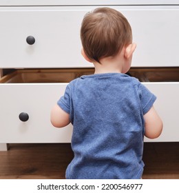 Toddler Baby Opens A Chest Of Drawers. Child Boy Reaches Into An Open Drawer Of A White Cabinet. Kid Age One Year