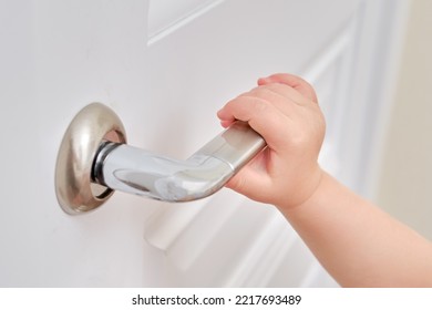 Toddler Baby Opens The Door Holding The Door Handle, Child Hand Close-up. White Wooden Door, Metal Door Handle And Baby Hand