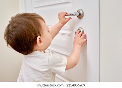 Toddler Baby Opens The Lock, Holding The Door Handle, Child Hand Close-up. White Wooden Door, Metal Door Handle And Baby Hand