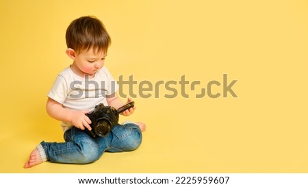 Toddler baby photographer with a camera on a studio yellow background. Happy child with photo equipment in a white t-shirt and blue jeans, copy space. Kid aged one year and four months