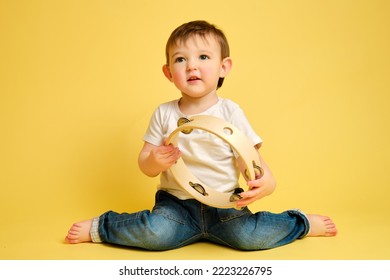 Toddler Baby Plays The Tambourine, A Child With A Percussion Musical Instrument On A Studio Yellow Background. Happy Child Musician Playing Hand Drum. Kid Aged One Year Four Months