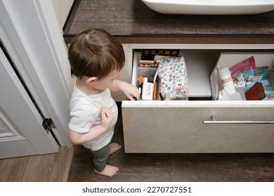 Toddler baby pulls out an item drawer in home bathroom  A small child explores the closet in the bathroom  Kid aged one year eight months