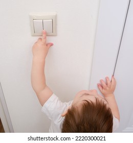 Toddler Baby Reaches For The Light Switch, Child Hand Turns On The Lamp Close-up. White Light Switch In Home Room And Baby Hand