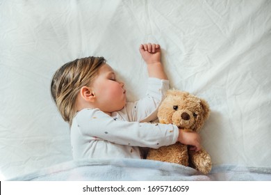 Toddler Baby Sleeping On White Sheets Hugging Soft Brown Teddy Bear. Children's Nap Time Or Bedtime. Child Sleep Deprivation. Child Sleep Routine And Schedule.