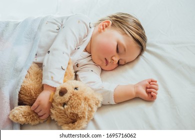 Toddler Baby Sleeping On White Sheets Hugging Soft Brown Teddy Bear. Children's Nap Time Or Bedtime. Child Sleep Deprivation. Child Sleep Routine And Schedule.