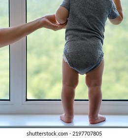 Toddler Baby Is Standing By The Window And Mother Hand Hold Him. Child At The Window Holding Hands On The Glass. Kid Age One Year