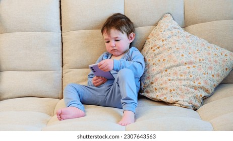 Toddler baby is tapping away on the mobile phone screen. The happy youngster is glued to the screen of their phone while seated comfortably on the couch. Kid aged about two years (one year ten months) - Powered by Shutterstock