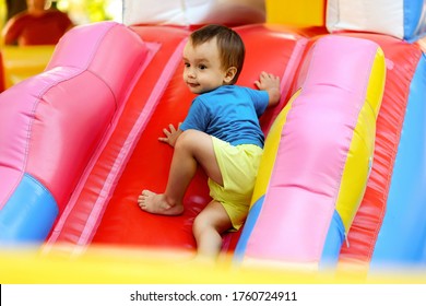 Toddler Boy In Blue Shirt And Yellow Shorts Climbing Up Slide On Bright Inflatable Bouncy Castle. Little Kid Half Turned On Bouncing Attraction At Playground, Maybe He Is Looking For Parents Attention