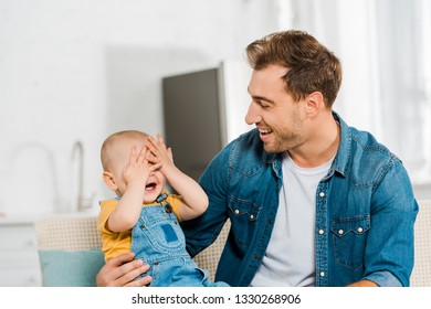 Toddler Boy Covering Eyes With Hands And Playing Peekaboo Game With Smiling Father At Home